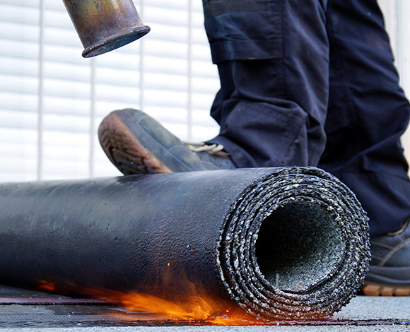 person applying heat to rolled roofing material with flames visible near the edge using safety gear for roofing installation and maintenance projects
