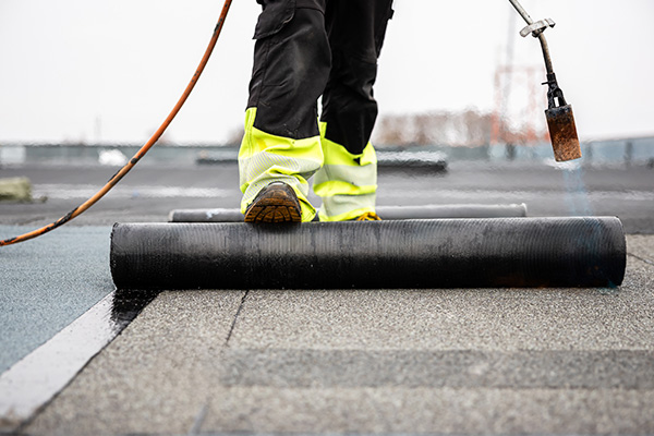 person applying roofing material with torch on a roll of roofing membrane on rooftop using safety gear for roof installation and repair 4 essential steps