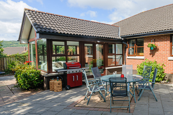 outdoor patio area with glass conservatory red barbecue grill and circular table surrounded by chairs featuring five bright plants and sunny sky