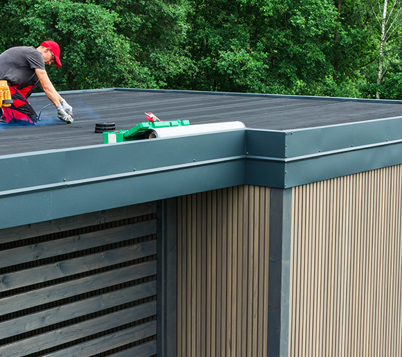 worker applying roofing material on a flat roof with green and white tools surrounded by trees for a 2 story building renovation