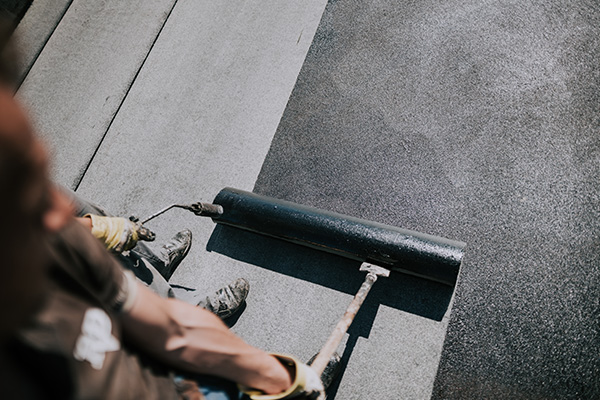 person applying black material with a roller on a surface showing multiple layers of asphalt five layers of coating application