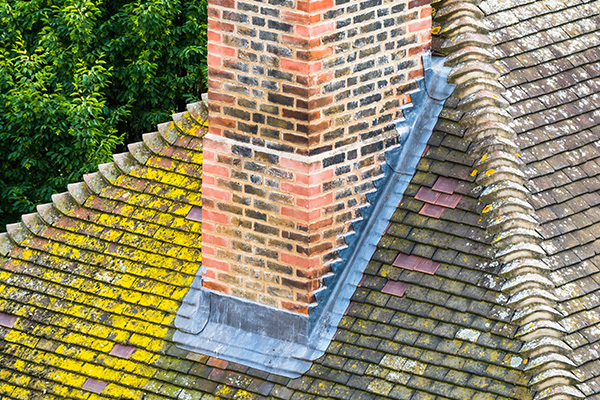 aerial view of a chimney on a sloped roof with moss and lichen growth showcasing roofing materials and the structure of the chimney in a weathered state maintaining two tones of tiles