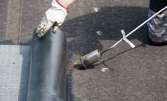 person using a torch to apply heat on a black roofing membrane during installation two layers of roofing material